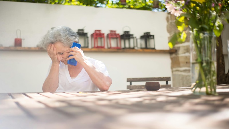 Person with gray hair sitting at an outdoor table, holding a blue phone to their ear. Behind them is a white wall with decorative lanterns on a wooden shelf and a vase of flowers to the right.